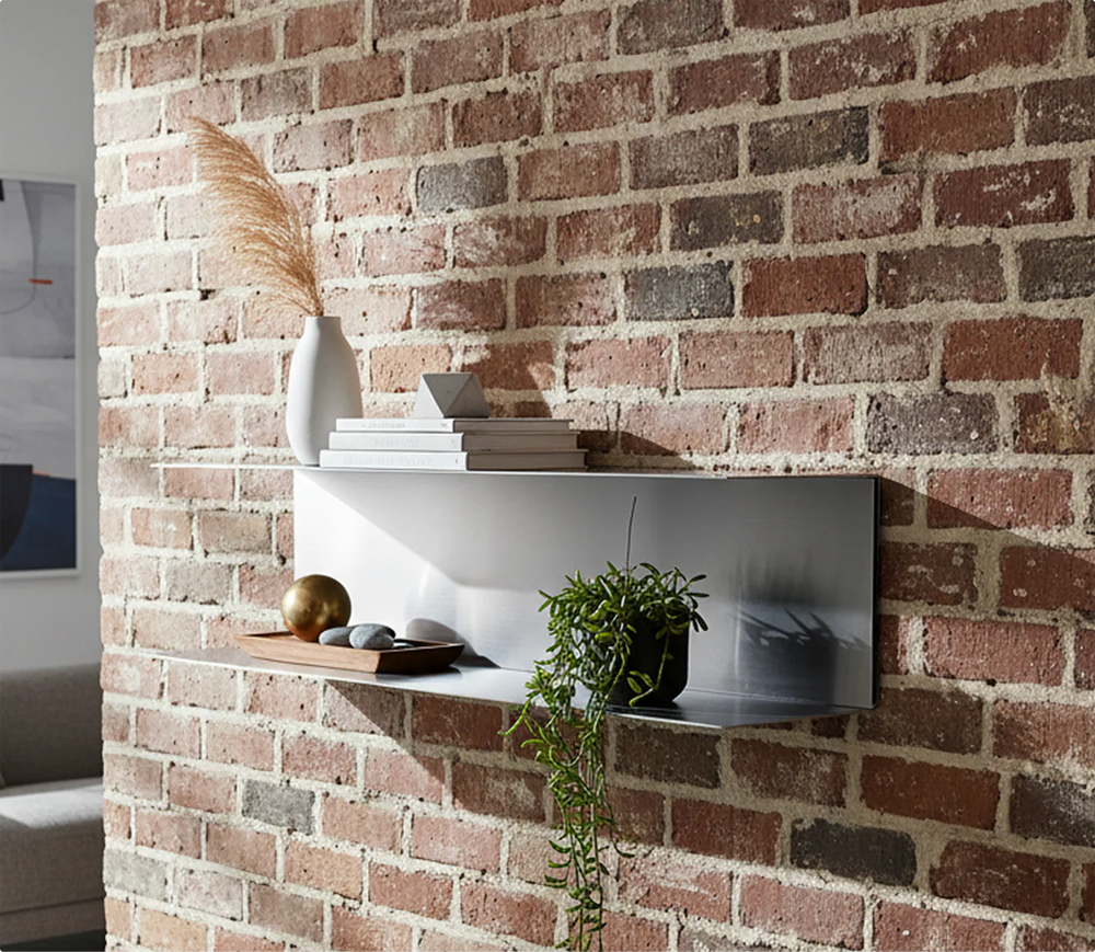 Handcrafted brushed metal two-tier floating shelf by Fig & Forge installed on a rustic red brick wall for a minimalist vintage design look